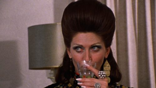 In a frame from George Romero's 1973 film SEASON OF THE WITCH, actress Jan White sips from a cocktail while rocking frosty eyeshadow and an astonishing beehive hairdo