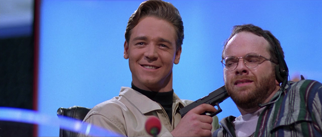 Russell Crowe menaces a hapless TV technician with a gun against a blue background in Virtuosity (1995)