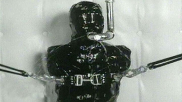 From the Broken movie by Nine Inch Nails, a black and white medium shot of someone restrained in head-to-toe latex and with a pipe going into their mouth, from which little jets of water are leaking